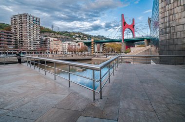 Modern müze mimarisi ile Bilbao nehir kenarındaki gezinti alanı, ikonik kırmızı kemer köprüsü ve şehir binaları bulutlu bir gökyüzünün altında, canlı bir seyahat sahnesinin altında nehre yansıdı.