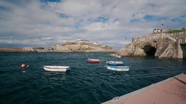 Küçük renkli balıkçı tekneleri Tapia de Casariego, Asturias limanındaki okyanus dalgalarında sallanıyor. Deniz feneri adası bulutlu bir günde manzaralı arka planda.