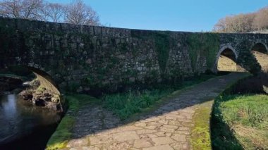 Galiçya, İspanya 'da bir nehrin üzerinde kemerleri olan antik taş köprü, güneşli bir günde Camino de Santiago' da yürüyen hacılar için bir dönüm noktası. Kamera kaldırım taşı yolu boyunca hareket ediyor.