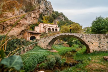 Bir derenin üzerindeki taş kemer köprüsünde duran kişi, güzel doğal bir manzarada uçurumun kenarına inşa edilmiş tarihi Tozalmoro inziva şapelini hayranlıkla izliyor.