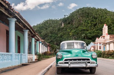 Geleneksel bir Küba köyünde renkli bir caddeye park edilmiş klasik yeşil araba, canlı mimarisi ve yemyeşil tropikal dağlar arka planı oluşturuyor.