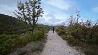 Çakıl taşlı bir yolda tek başına yürüyen, yemyeşil tepelerle çevrili, açık havada doğa ve yalnızlığı kucaklayan, bulutlu bir gökyüzünün altında, mavi benekli, doğa ve yalnızlık dolu bir yürüyüş macerası.