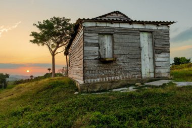 Gün batımında vadiye bakan çimenli bir tepenin üzerinde yıpranmış ahşap bir ev ılık altın bir ışık kırsal Küba kırsalını ve huzurlu gökyüzünü renklendiriyor.