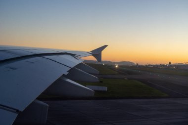 Gün batımında bir uçak kanadından gelen hava manzarası, pisti ve uzak havaalanı ışıklarını gösteriyor. Gökyüzü turuncudan maviliğe geçerek huzurlu bir yolculuk atmosferi yaratıyor..
