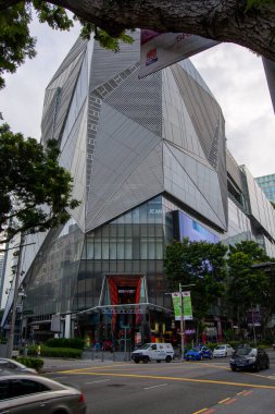 Benzersiz geometrik tasarımı olan, cam ve metal elementlere sahip modern bir mimari bina, ön planda ağaçlar ve araçlarla kentsel bir ortamda yer alıyor.