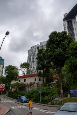 Modern binalar ve yeşilliklerin yer aldığı bulutlu bir şehir manzarası, sokakta yürüyen bir kişi...