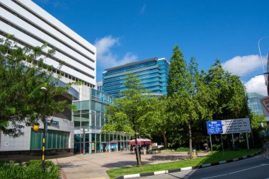 Ticari binalar, yemyeşil alanlar ve açık mavi gökyüzünün yer aldığı modern bir şehir manzarası.