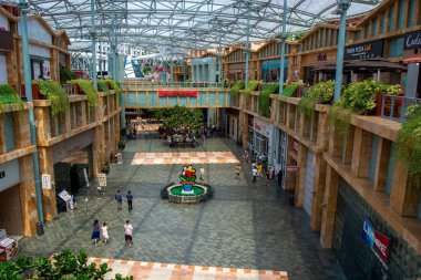Modern tasarıma sahip geniş bir alışveriş merkezi, açık alanlar, yeşillik ve ziyaretçilerle dolu merkezi bir dekoratif özelliğe sahip.