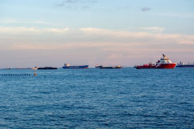 Gün batımında denizde birkaç geminin bulunduğu bir liman manzarası. Kargo gemileri ve kırmızı bir kurtarma gemisi var.