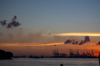Gökyüzünde canlı renkler ve endüstriyel yapıların siluetleri olan vinçler ve teknelerle dolu bir limanın üzerinde manzaralı bir günbatımı.