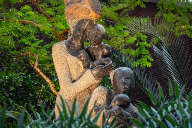 Yeşilliklerle çevrili iki çocuğu tutan bir annenin taş heykeli.