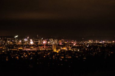 Geceleri gökyüzüne bakan bir şehrin panoramik görüntüsü, renkli havai fişeklerle aydınlatılıyor, ön planda ve arka planda binalar ve ışıklar sergileniyor.