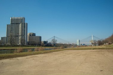 Modern binalarla dolu bir nehir manzarası ve berrak mavi gökyüzünün altında kablo destekli bir köprü.