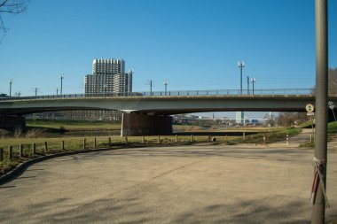 Arka planda şehir silueti, açık mavi gökyüzü ve ön planda yeşil çimenler olan bir nehir üzerinden geçen modern bir köprü.