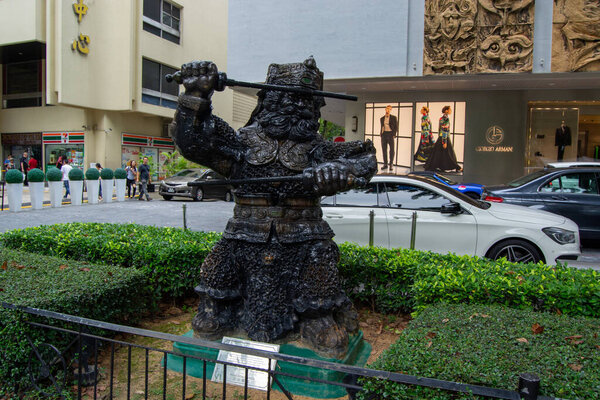 A large black statue of a warrior holding two swords, surrounded by greenery and modern buildings