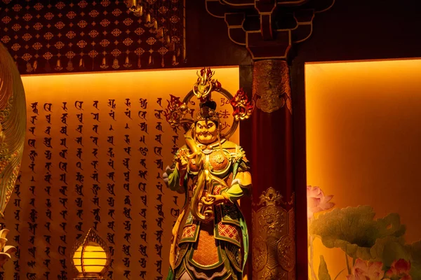 Tapınak ortamındaki renkli bir tanrı heykeli. Arkasında geleneksel Asya kaligrafisi ve sıcak ışıkla aydınlatılmış.
