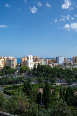 A panoramic view of a coastal city featuring modern buildings, lush greenery, and a clear blue sky