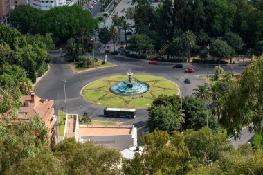 Merkezinde bir çeşme olan bir kavşağın havadan görünüşü, yeşillik ve palmiye ağaçları ile çevrili, yolda arabalarla şehir merkezi.