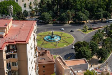 Merkezinde yeşil çimenler ve palmiyelerle çevrili bir çeşme olan döner kavşağın havadan görünüşü. Binalarla çevrili kentsel ortam.