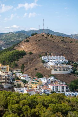 Yeşillik ve dağlarla çevrili yerleşim binaları ve iletişim kuleleri olan bir tepenin manzarası mavi bir gökyüzünün altında..