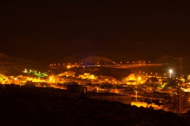 Arka planda tepeler olan sokak lambaları ve binalarla aydınlatılmış küçük bir kasabanın gece manzarası.