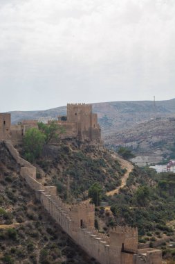 Kayalık arazi ve yeşilliklerle çevrili, bulutlu bir gökyüzünün altında uzanan dolambaçlı bir yolu olan tarihi bir şato.