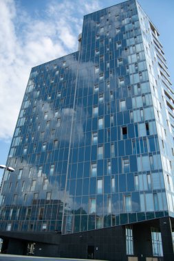 Yansıtıcı cam cepheli, gökyüzü ve bulutların yansımalarını gösteren, mavi gökyüzüne karşı inşa edilmiş modern bir gökdelen.