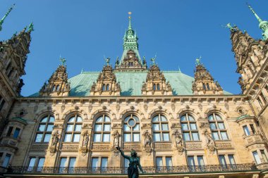 Açık mavi gökyüzünün altında yeşil bakır çatı ve heykellerin yer aldığı tarihi bir binanın alçak açılı görüntüsü.