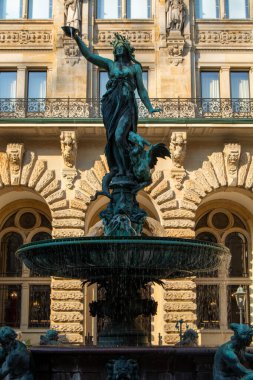 Elinde bardak tutan bir kadın heykeli olan dekoratif bir çeşme. Arka planda su ve karmaşık mimari detaylarla çevrili.