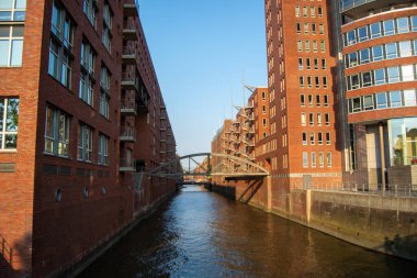 Kırmızı tuğlalı binalarla çevrili bir kanal manzarası, açık mavi bir gökyüzünün altında, suyun üzerinden geçen bir köprü.