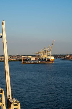Bir liman manzarası, vinçler ve kargo gemisi, mavi gökyüzü ve sakin su.