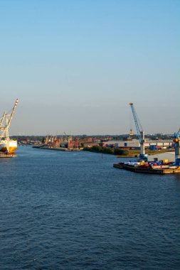 Açık mavi gökyüzünün altında, kıyılar boyunca vinçleri ve konteynırları olan bir nehir manzarası.