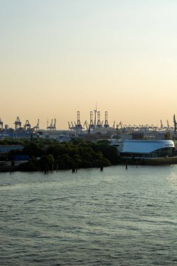 Gün batımında bir liman manzarası, rıhtım boyunca vinçler ve endüstriyel binalar var.