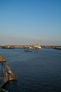 Arka planda rıhtım ve sanayi bölgesi olan bir nehir manzarası, açık mavi gökyüzünün altında vinçler ve bir mavna var.