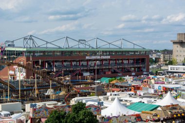 Ön planda panayır olan, renkli çadırların ve bineklerin yer aldığı değirmenci-stadyum manzarası