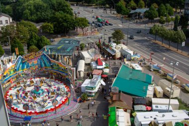 Canlı bir eğlence parkının havadan görünüşü. Renkli gezintileri ve yemek tezgahları var. Ağaçlarla çevrili ve araçları olan bir yol.
