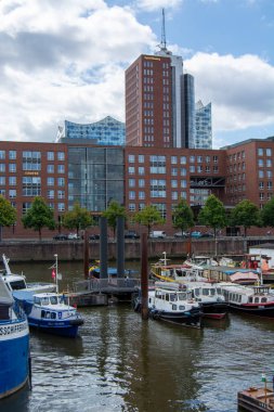 Parçalı bulutlu bir gökyüzünün altında modern binalar ve ağaçlarla çevrili çeşitli teknelerle dolu bir limanın manzarası.