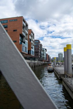 Nehir boyunca çağdaş binaların olduğu, teknelerin kenetlendiği ve bulutlu bir gökyüzünün olduğu modern bir şehir sahnesi.