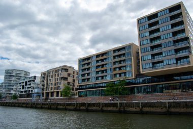 Nehir boyunca uzanan modern rıhtım binaları, cam cepheleri ve yeşillikler, bulutlu bir gökyüzünün altında.