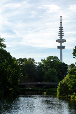 Arka planda yemyeşil ağaçlar ve açık mavi gökyüzü ile çevrili uzun bir iletişim kulesinin bulunduğu gölü olan bir parkın manzarası.