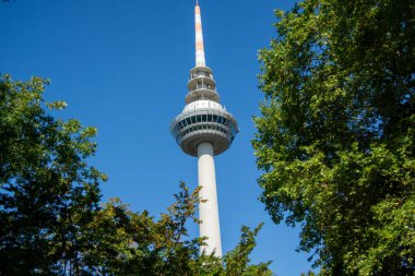 Açık mavi gökyüzünün altında yeşil ağaçlarla çevrili yüksek bir iletişim kulesi.