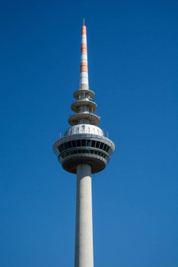 Silindirik tabanlı ve sivri uçlu, açık mavi gökyüzüne karşı kurulmuş uzun bir telekomünikasyon kulesi.
