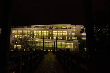 Gece aydınlatılan modern bir bina, ahşap bir geçitten görülüyor, büyük pencereler sıcak ışığı yansıtıyor.