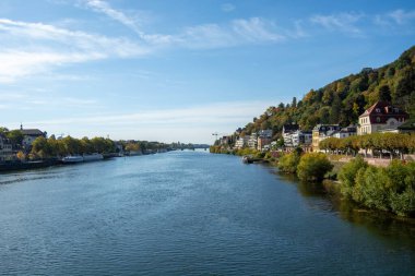 Bereketli yeşilliklerle ve açık mavi gökyüzünün altındaki binalarla çevrili bir nehrin manzarası.