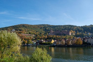 Yeşil tepeler ve sonbahar yapraklarıyla çevrili, kıyısında bir kasaba olan bir nehrin manzarası. Tepenin yamacında açık mavi bir gökyüzünün altında tarihi bir kale görülebilir..
