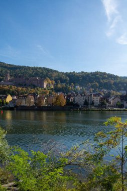 Sonbahar renklerinde tepeler ve ağaçlarla çevrili, açık mavi gökyüzünün altında kıyısı olan bir nehrin manzarası.