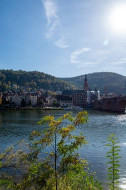 Renkli binaları, bir köprüsü ve berrak mavi gökyüzünün altındaki yemyeşil tepeleri olan nehir kenarı manzarası.