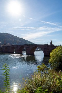 Nehir üzerindeki tarihi bir taş köprünün manzarası. Güneş ışığı suya ve tepelere ve ağaçlara yansıyor..