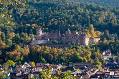 Sonbahar yeşillikleriyle çevrili tarihi bir şatonun panoramik manzarası, ön planda bir kasaba ve arka planda yemyeşil tepeler.