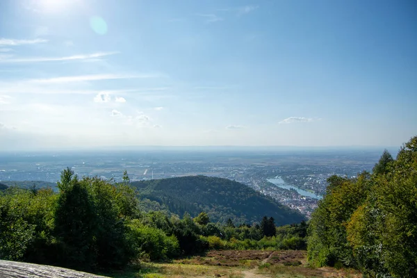 Tepenin tepesinden panoramik bir manzara, önplanda yemyeşil ağaçlar, şehirde kıvrılan bir nehir, ve dağınık bulutlu açık mavi bir gökyüzü.